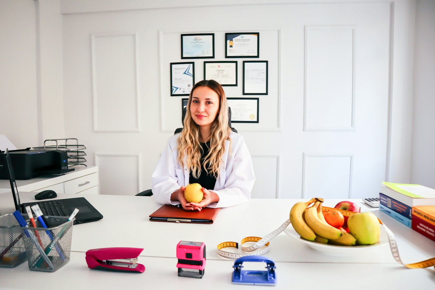Young woman in lab coat holds green apple at office desk.